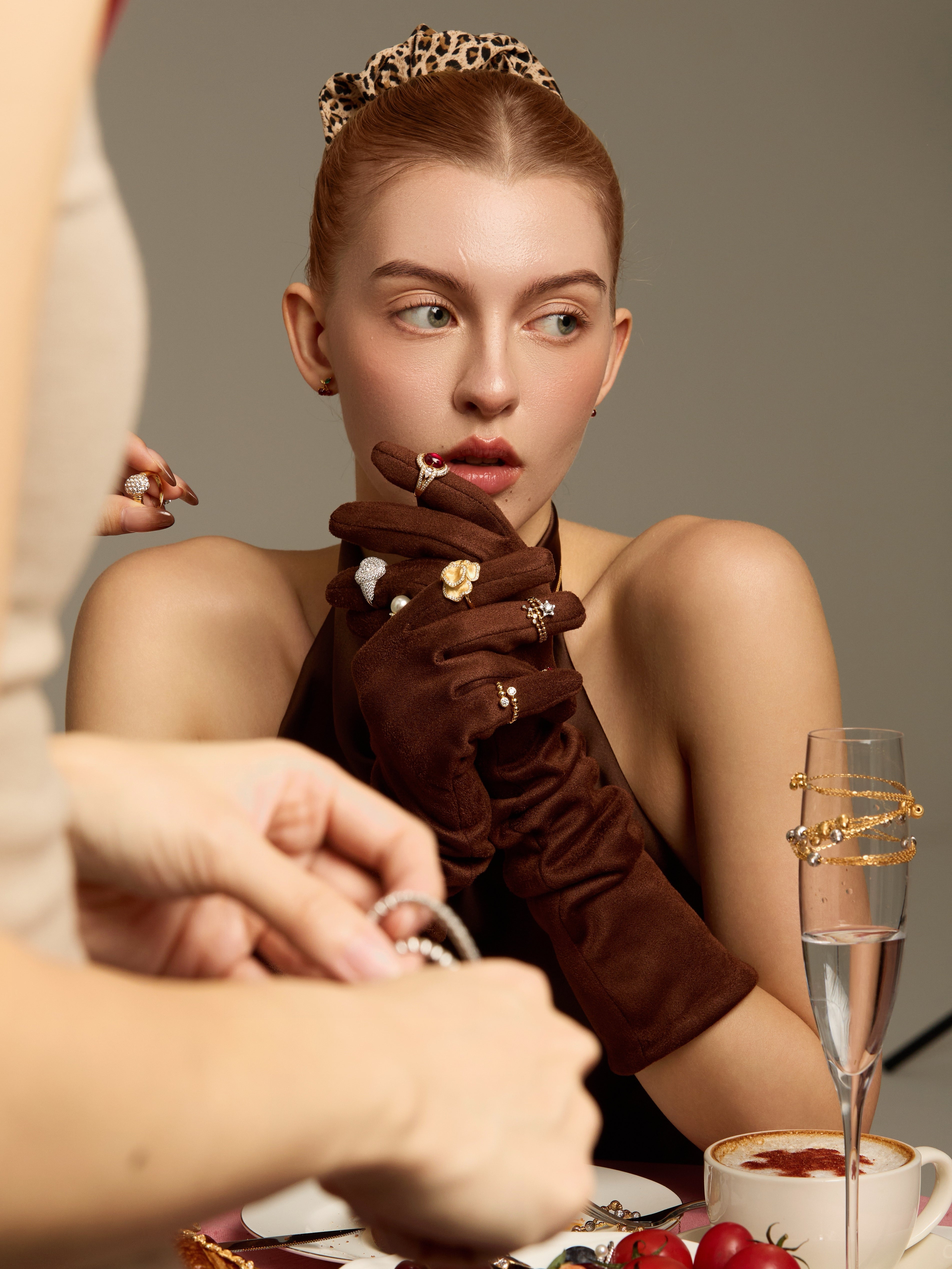 Model in a black evening look and brown gloves wearing multiple statement rings at a dinner table.