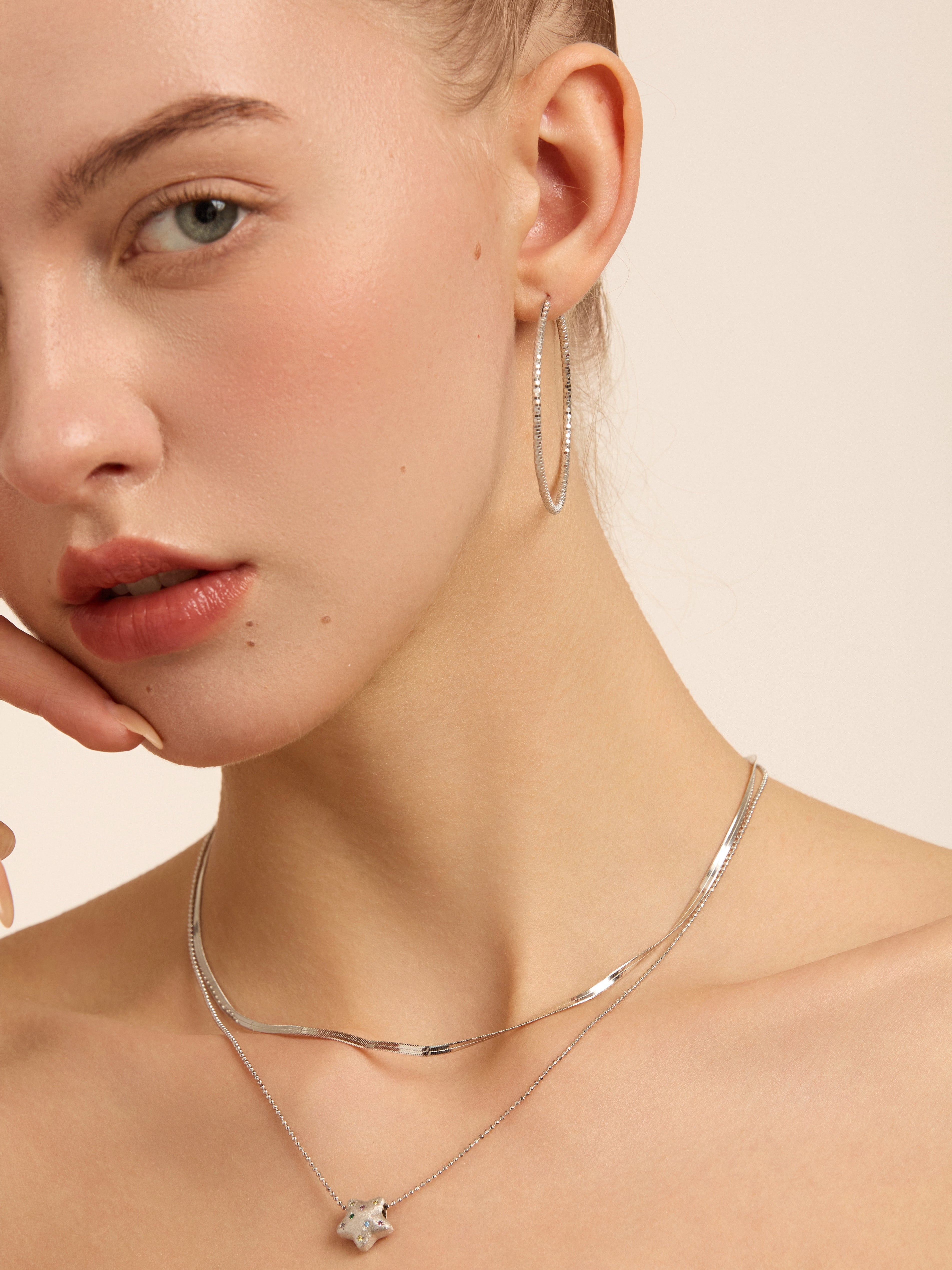 Model wearing a large silver-tone hoop with layered silver necklaces and a star pendant.